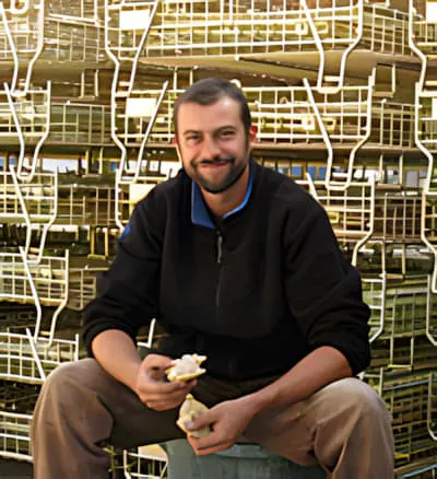 Un homme souriant en vêtements sombres assis parmi des caisses métalliques de bouteilles de vin empilées pendant le processus de mise en bouteilles.