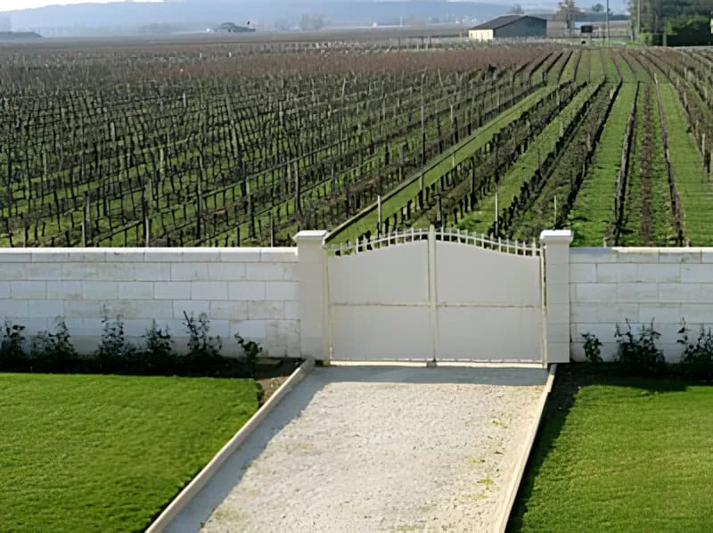 Un portail d'entrée formel avec des piliers en pierre blanche menant dans un vignoble, avec des rangées nettes de vignes s'étendant au loin sous des cieux clairs.