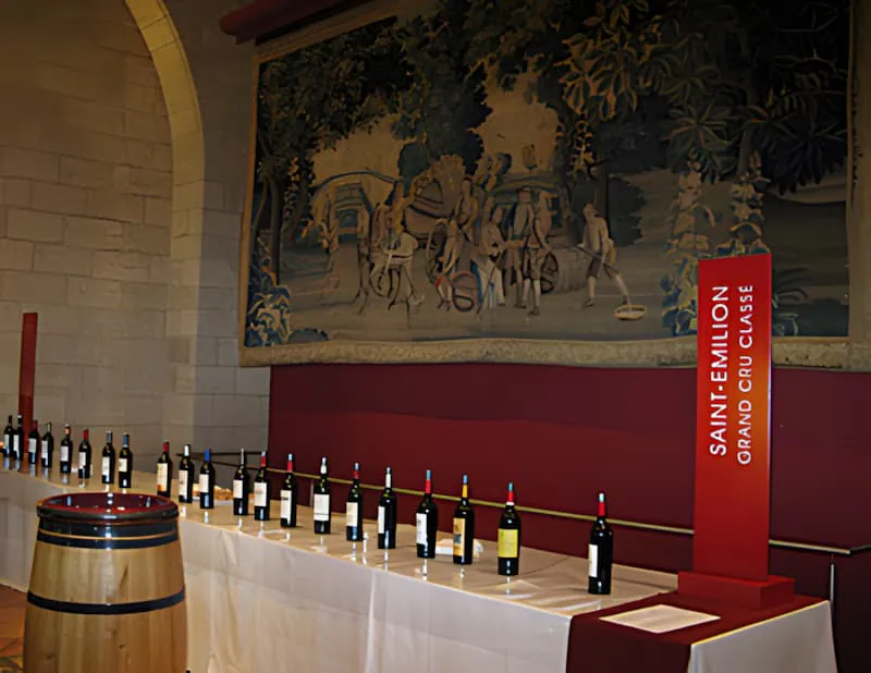 Une longue table dressée pour une dégustation de vin, avec de nombreuses bouteilles de vin rouge et verres, dans une grande salle avec une tapisserie au mur.
