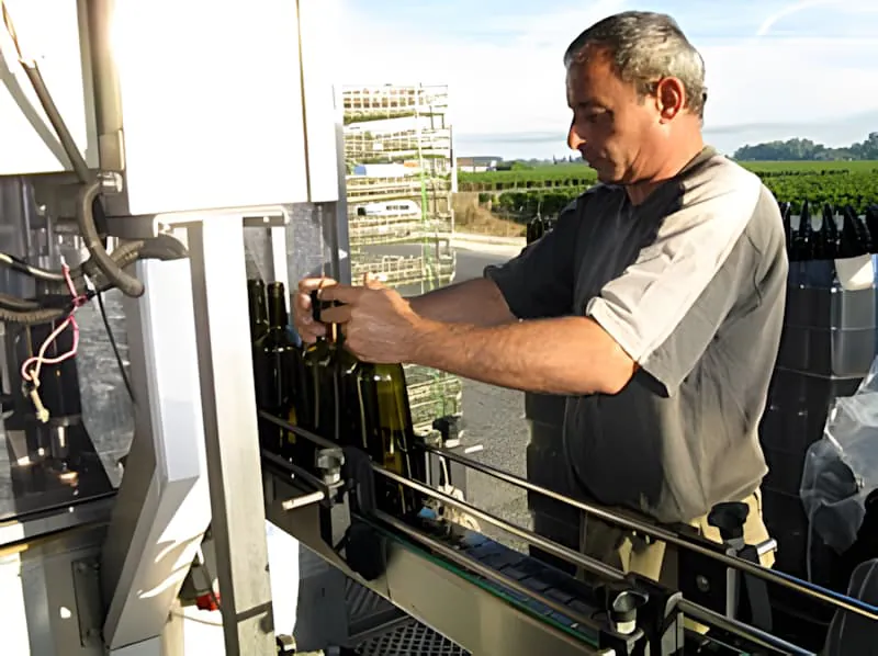 Un homme faisant fonctionner une machine de mise en bouteille, chargeant des bouteilles vides dans l'équipement tout en portant des vêtements de travail décontractés et une casquette.