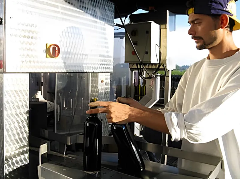Un ouvrier en casquette plaçant des bouteilles de vin remplies dans des caisses de stockage métalliques sur la ligne de mise en bouteille, montrant le côté final du processus.
