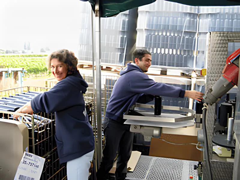 Un ouvrier de vignoble, souriant, charge des bouteilles de vin vides dans des cages métalliques sur un tapis roulant au Château Plaisance.
