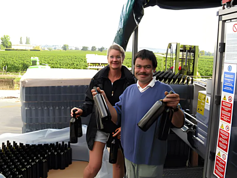 Un ouvrier de vignoble, souriant, charge des bouteilles de vin vides dans des cages métalliques sur un tapis roulant au Château Plaisance.