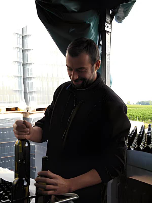 Un ouvrier de vignoble, souriant, charge des bouteilles de vin vides dans des cages métalliques sur un tapis roulant au Château Plaisance.
